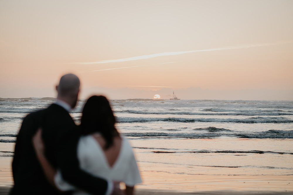 The newlyweds looking out into the Pacific Ocean, with the setting sun and a small boat in the distance at Cannon Beach. 