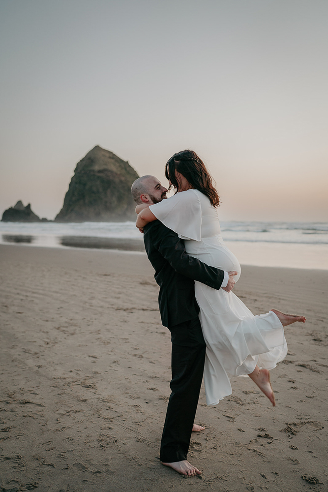 The groom carrying the bride on the sand with Haystack Rock in the backround at Cannon Beach. 
