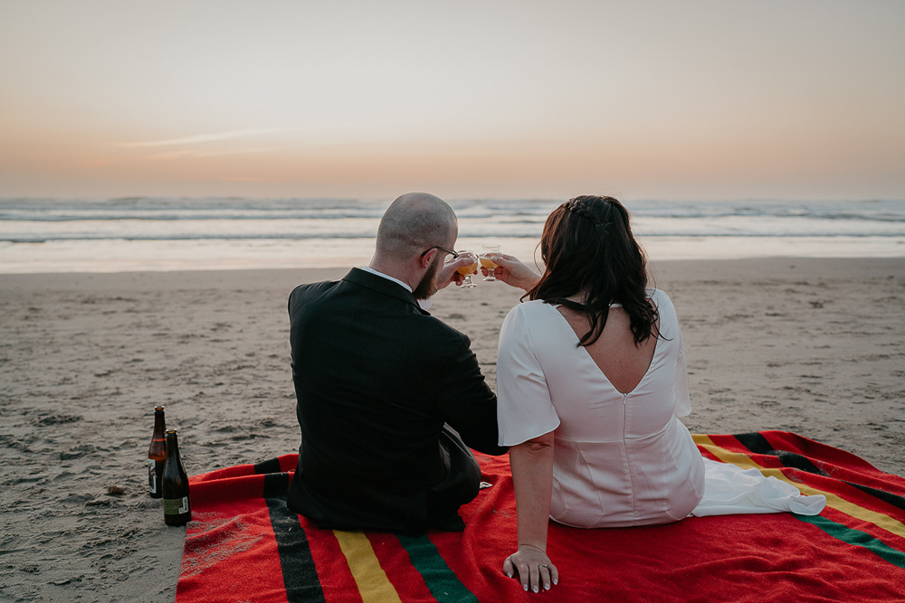 The newlyweds sitting on a Pendleton blanket cheersing while watching the sunset at Cannon Beach. 