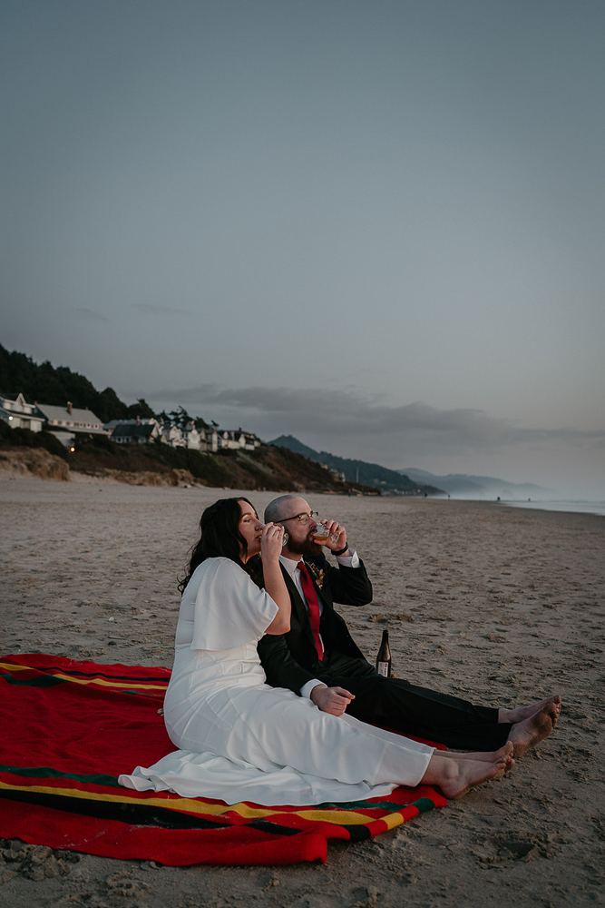 The newlyweds sitting on a Pendleton blanket drinking beer while watching the sunset at Cannon Beach. 