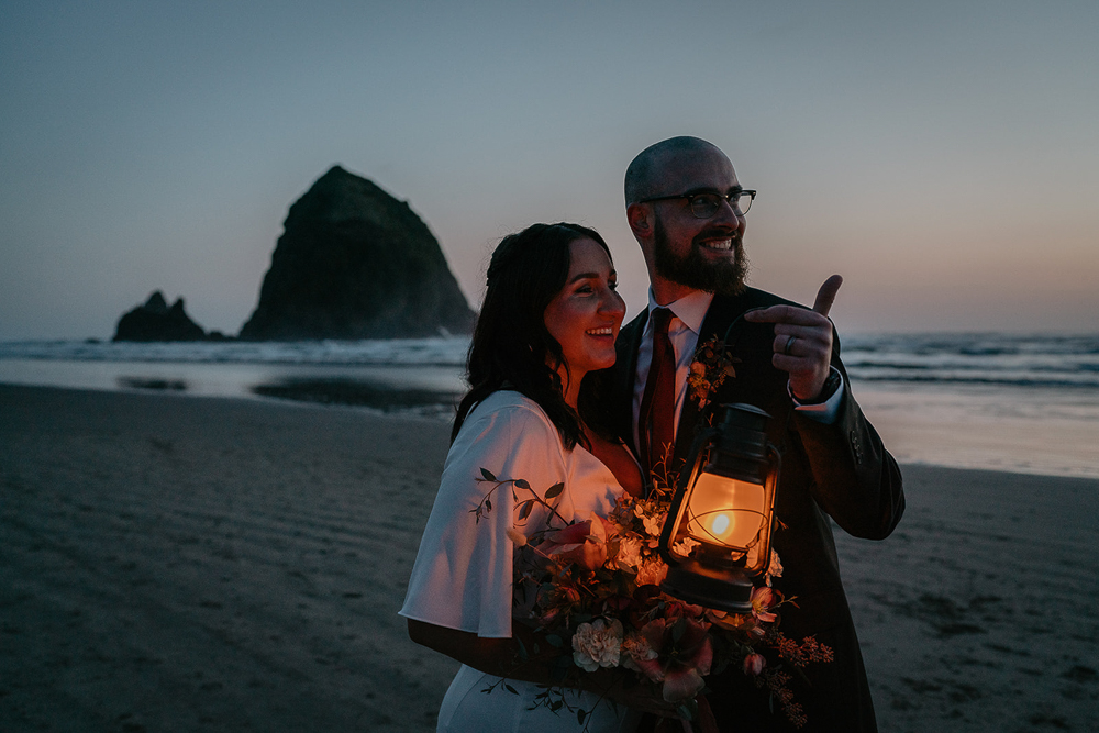 Newlyweds lit up by the a lantern and holding a flower bouquet with Haystack Rock in the background at Cannon Beach. 