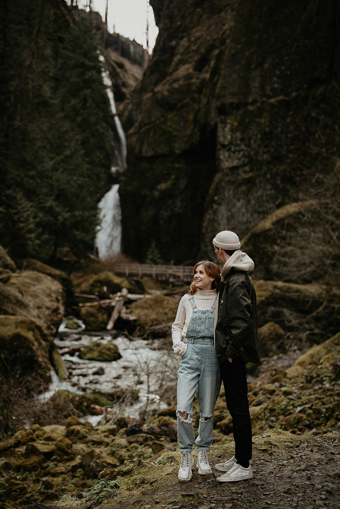 A couple standing side by side during the engagement session with a river and waterfall behind them at the Columbia River Gorge. 