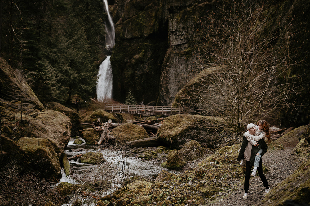 A man carrying his partner while walking on a trail with a bridge and waterfall in the background at the Columbia River Gorge. 