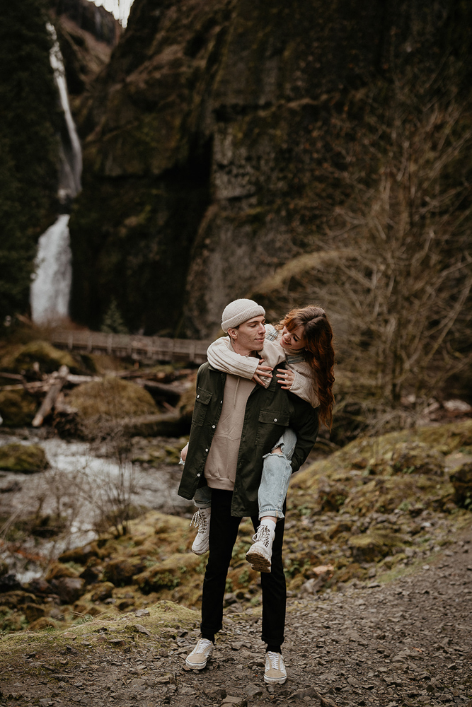 A man carrying his partner during their engagement session with a river and waterfall in the background at the  Columbia River Gorge. 