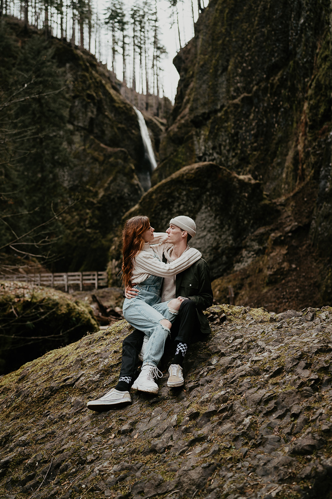 A couple siting on a mossy rock and hugging with a bridge and waterfall in the background at the Columbia River Gorge. 