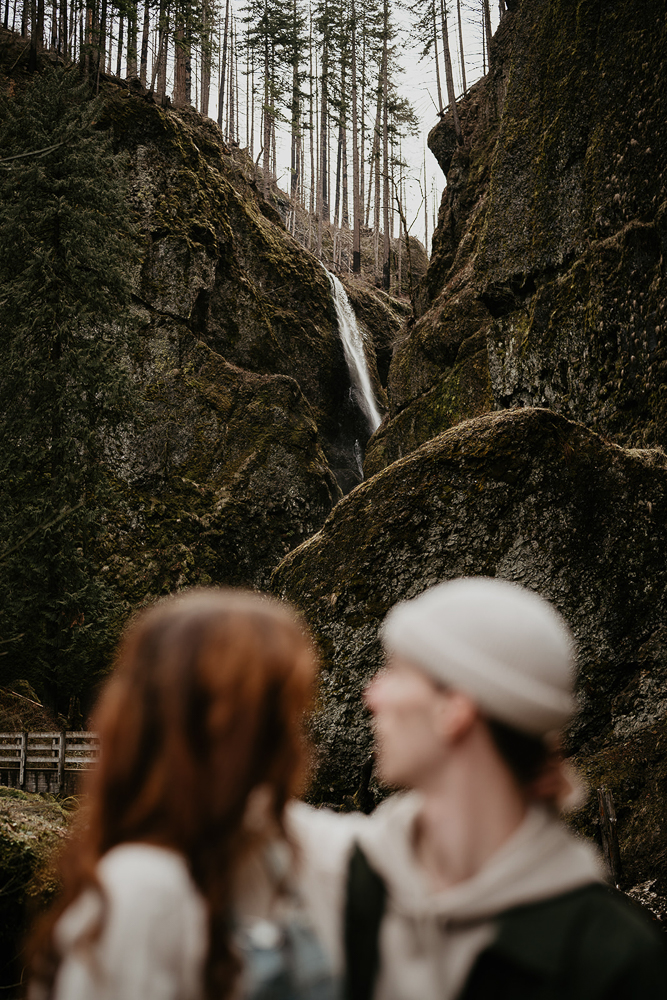 A blurry photo of a couple with a bridge, mossy boulders, and a waterfall in the background at the Columbia River Gorge. 
