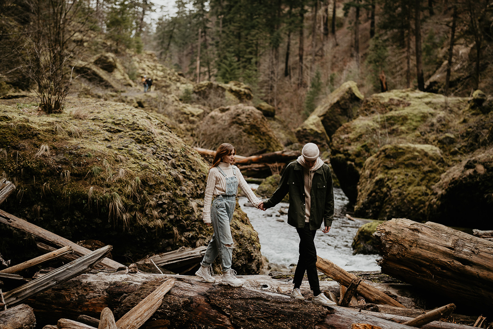 A couple holding hands and walking over a natural bridge with mossy rock and trees in the background at the Columbia River Gorge. 