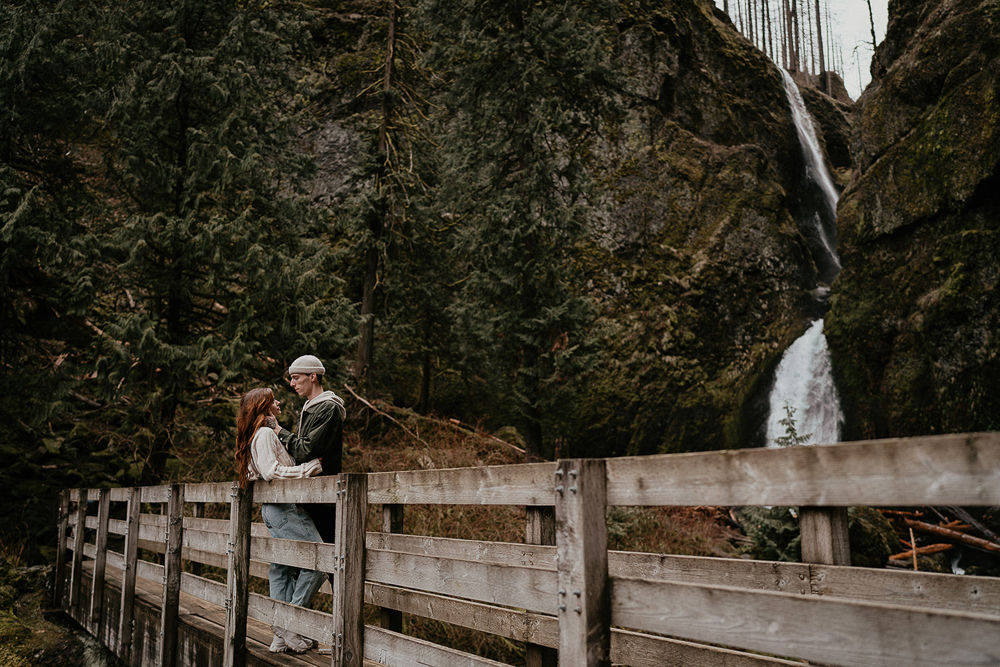 A couple leaning against a bridge rail with a waterfall and trees in the background at the Columbia River Gorge. 