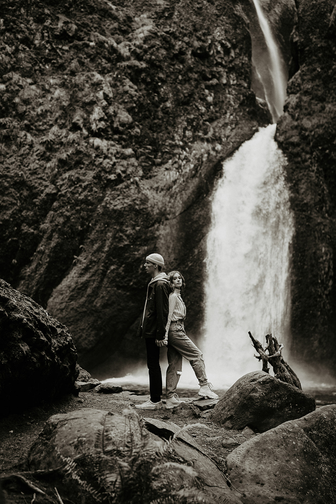 A black and white photo of a couple holding hands and standing back to back with a waterfall and mossy boulders in the background at the Columbia River Gorge. 