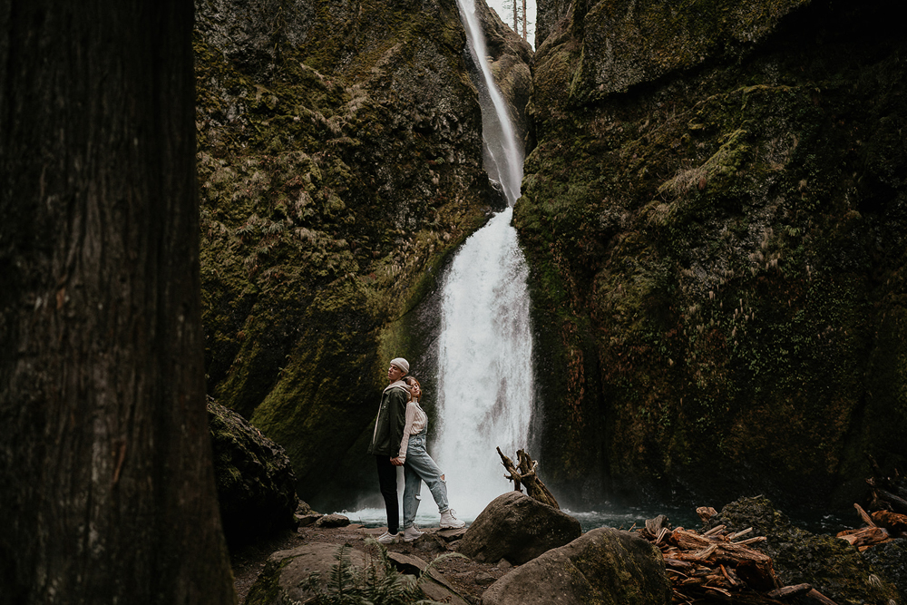 A couple holding hands and standing back to back with a waterfall and mossy boulders in the background at the Columbia River Gorge. 