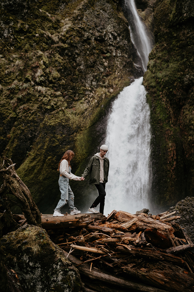 A couple holding hands and walking over a natural dam with a waterfall in the background at the Columbia River Gorge. 