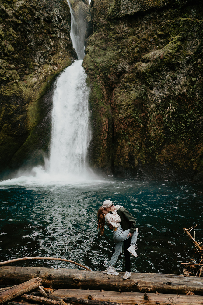 A couple standing on a log with a waterfall and river in the background at the Columbia River Gorge. 