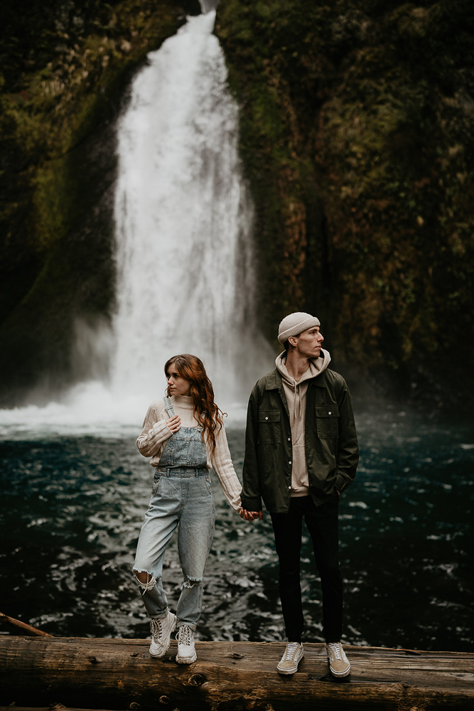 A couple holding hands while standing on a log with a waterfall in the background at the Columbia River Gorge. 