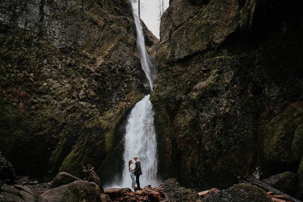 A couple holding each other while standing on a pile of logs with a waterfall in the background at the Columbia River Gorge. 