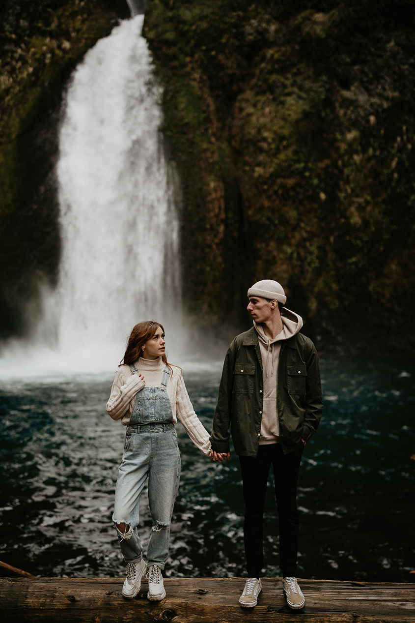 A couple holding hands while standing on a log with a waterfall in the background at the Columbia River Gorge.