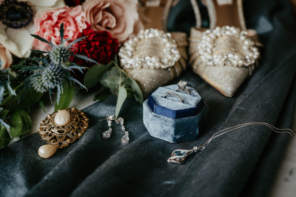 Blue velvet wedding ring box sitting on top of a blue cloth with sparkly heels and wedding flowers
