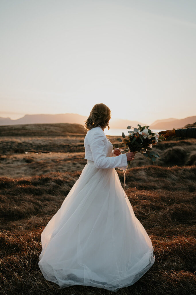 A bride in a white dress holding a bouquet of flowers, standing in a field as the sun sets in Iceland. 