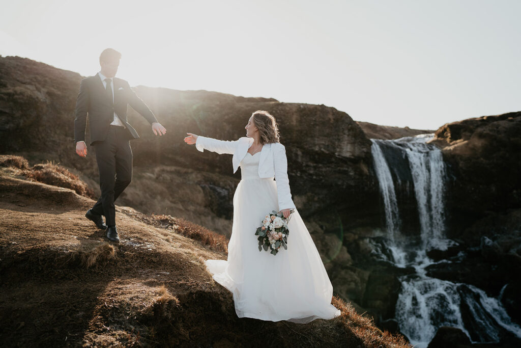 A bride and groom reaching out for each other with a waterfall raging in the background. 