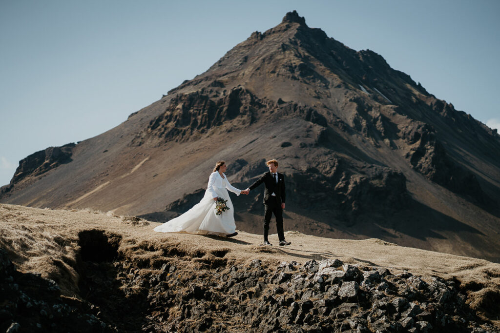 A bride and groom holding hands with a large Icelandic mountain looms in the distance. 