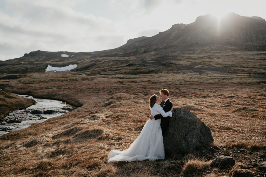 A bride and groom hugging and looking lovingly into each other's eyes while standing in an Icelandic field and with a river bubbling past them. 