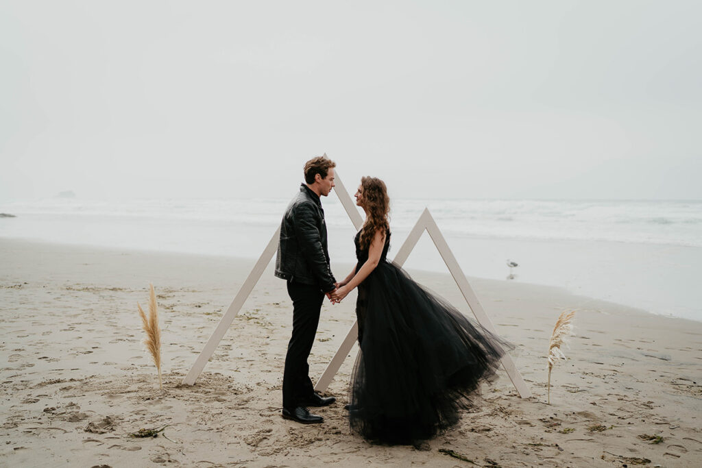 A couple dressed in all black holding hands in front of two large triangles on the Oregon coast. 
