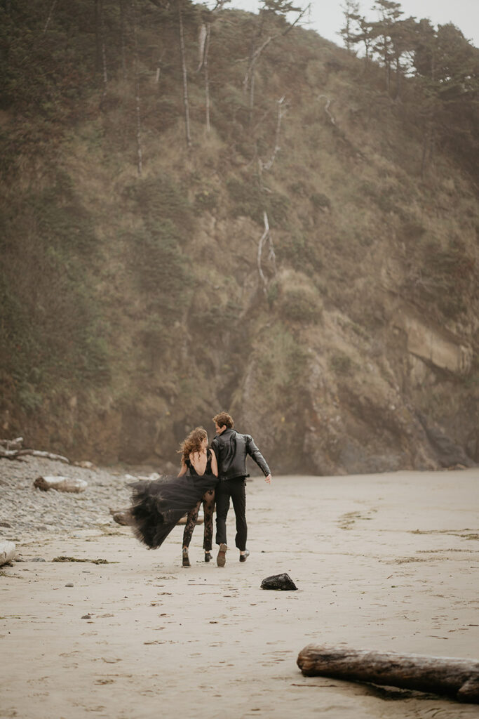 a couple dressed in all black running away from the camera on the beach. 