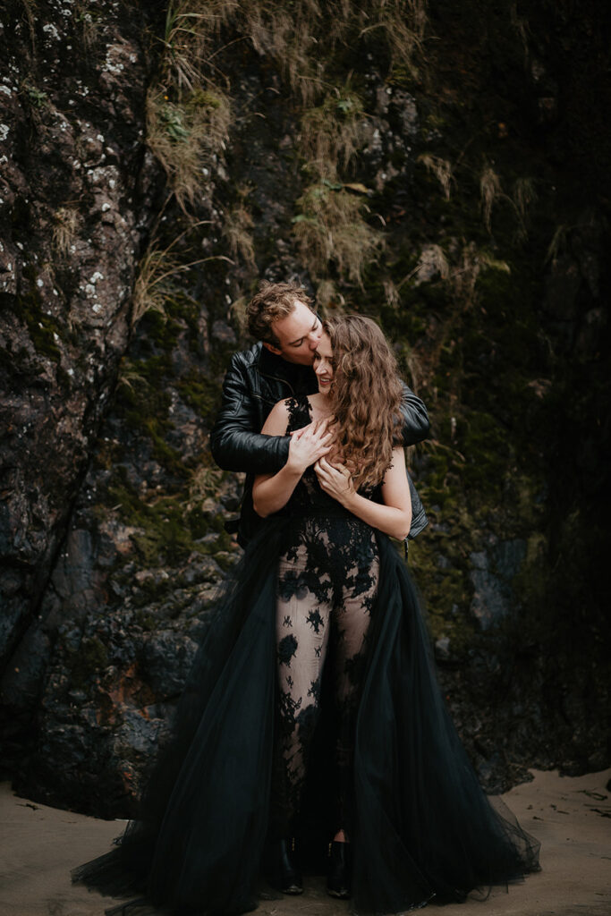 A couple dressed in all black kissing in front of some mossy rocks. 
