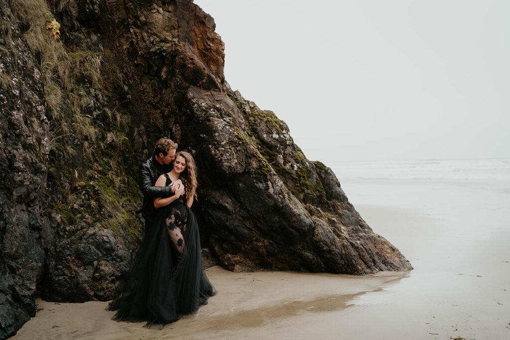 A couple dressed in all black hugging next to mossy rocks on the Oregon coast. 