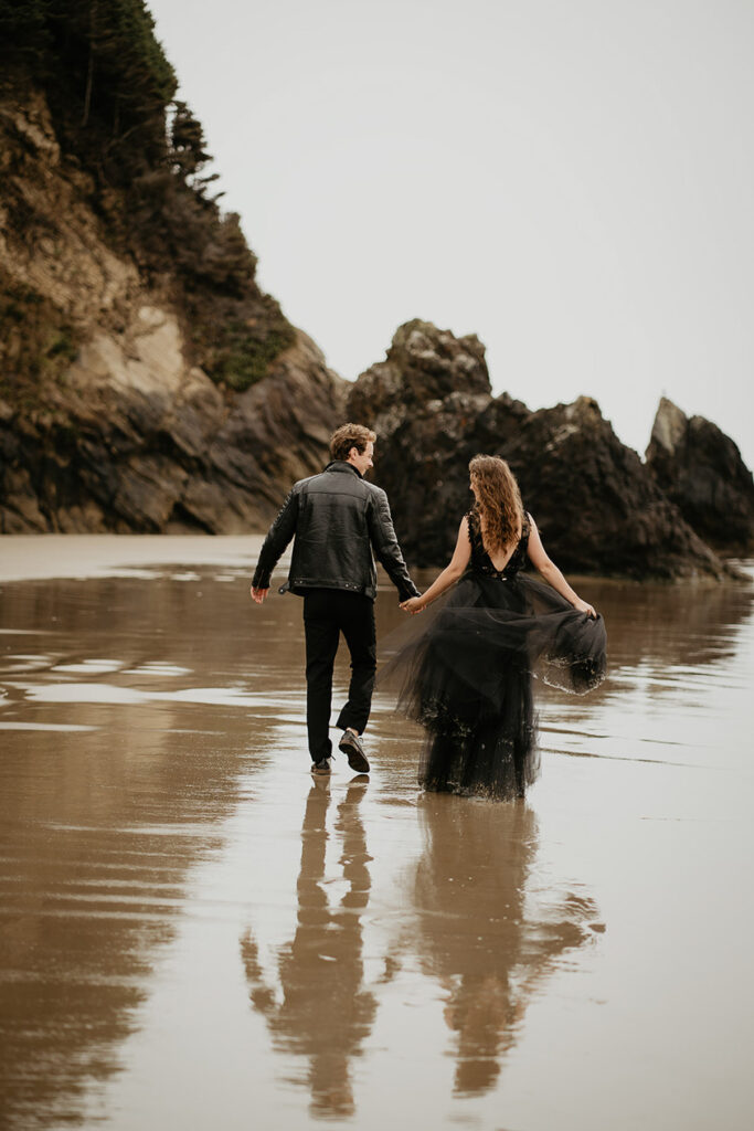 A couple dressed in all black walking away from the camera on the beach. 