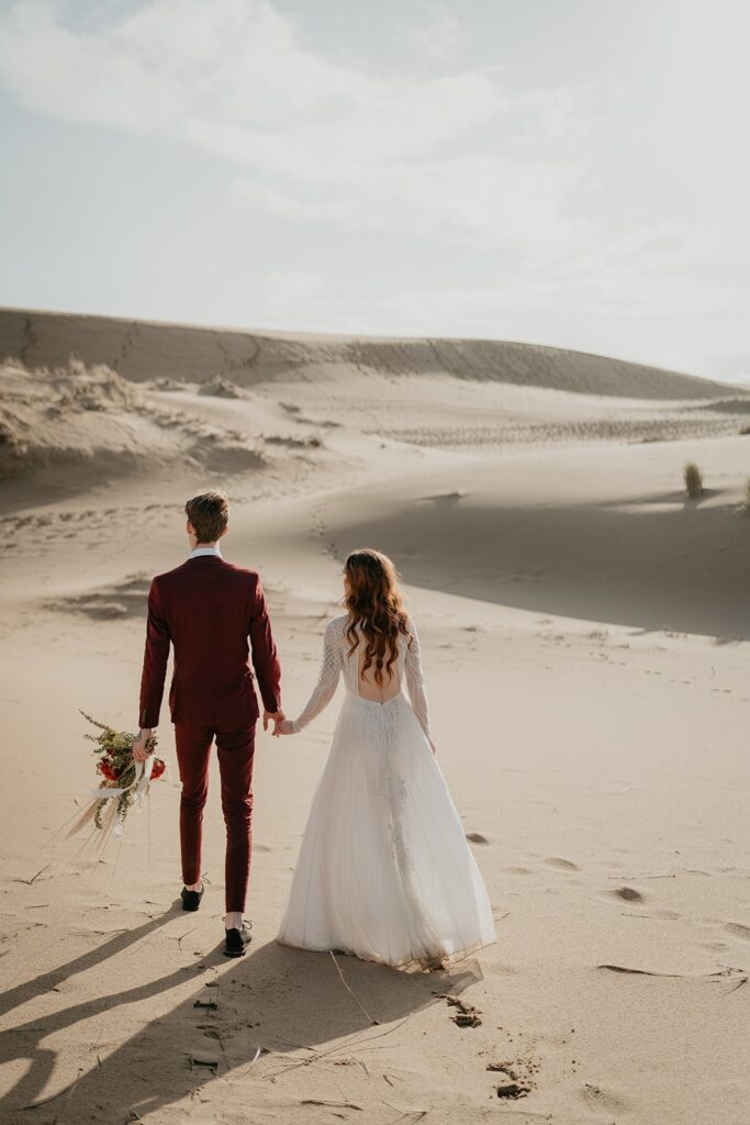 Bride and groom hold hands while walking across the sand dunes in Oregon