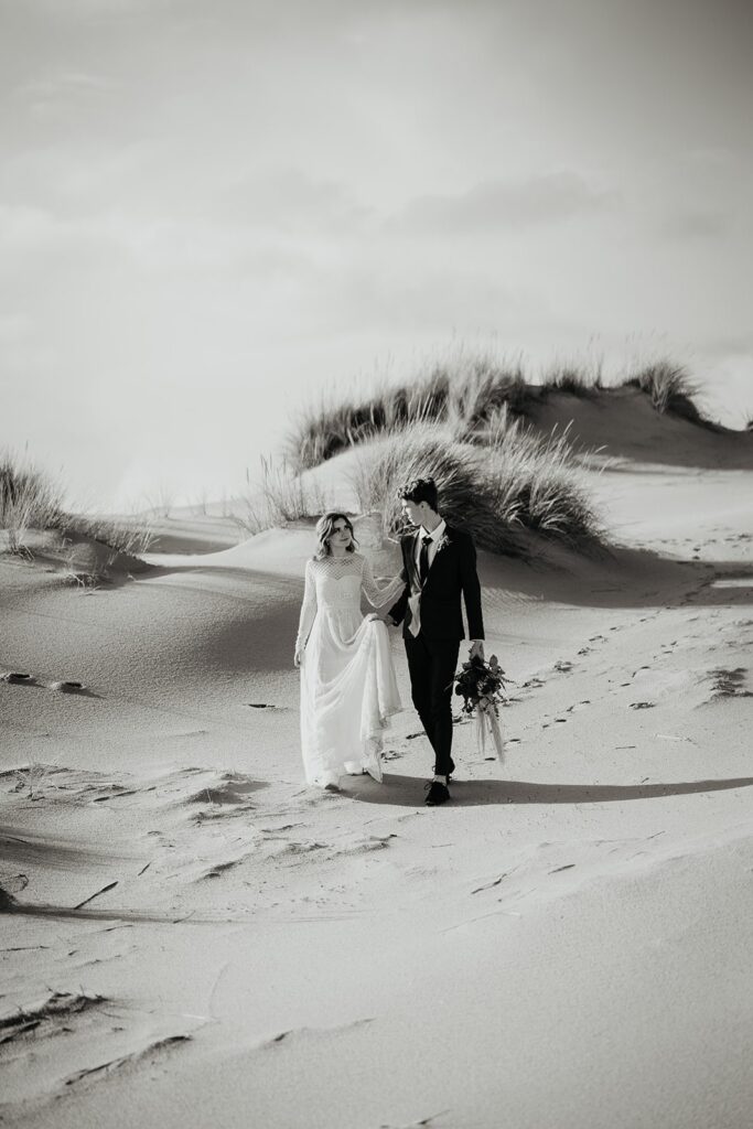 Bride and groom hold hands while walking across the sand dunes in Oregon