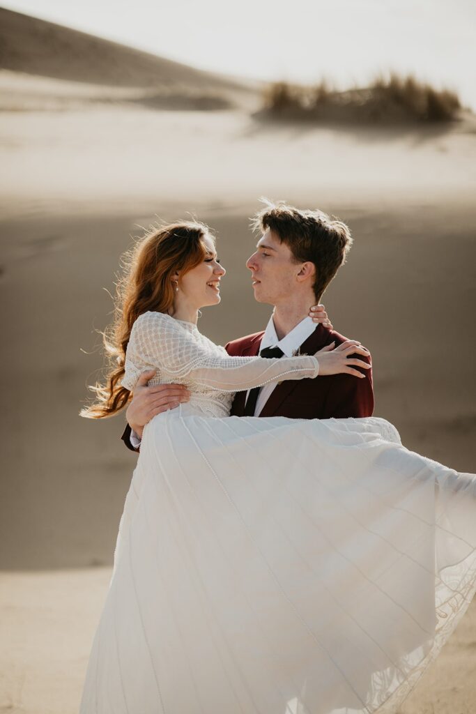 Groom carries bride during elopement photos on the sand dunes in Oregon