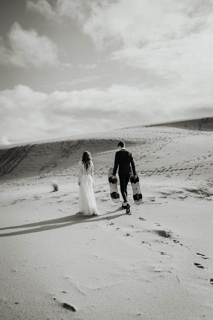 Bride and groom walking across the sand dunes while holding sand boards in Oregon for their adventure elopement