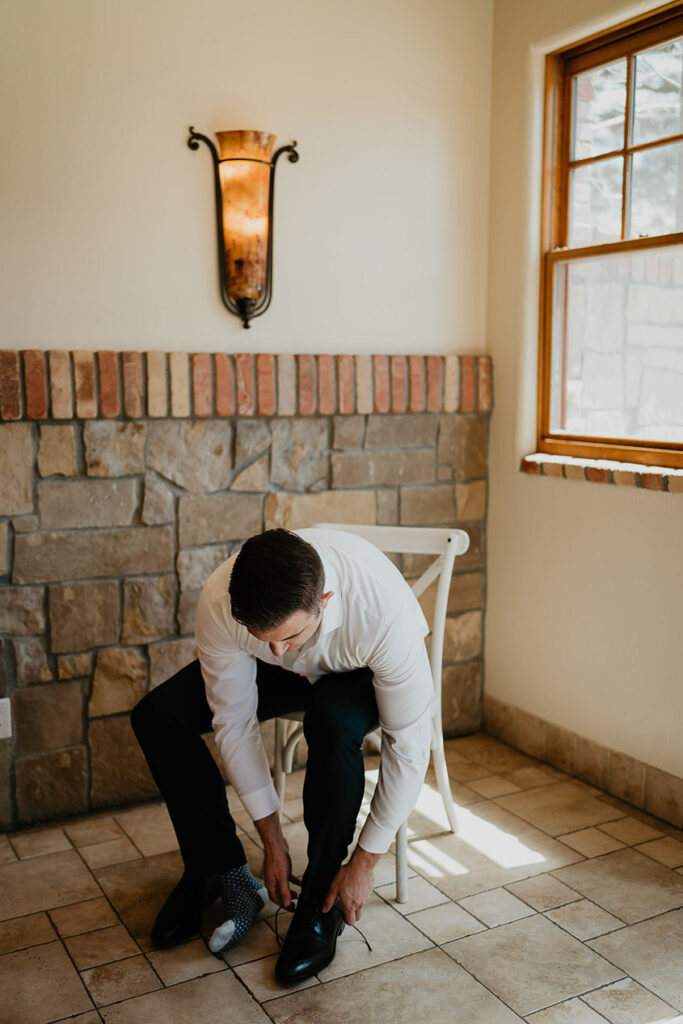 The groom sitting in a rustic room tying his shoe. 