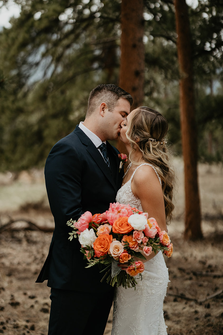 The bride, holding a rose bouquet, and groom kissing in the forest in Estes Park before their wedding.