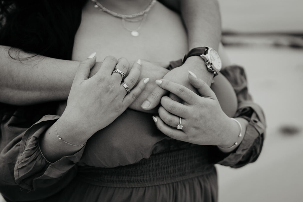 A black and white closeup on the boyfriend holding his girlfriend. You can see her rings and his watch clearly. 