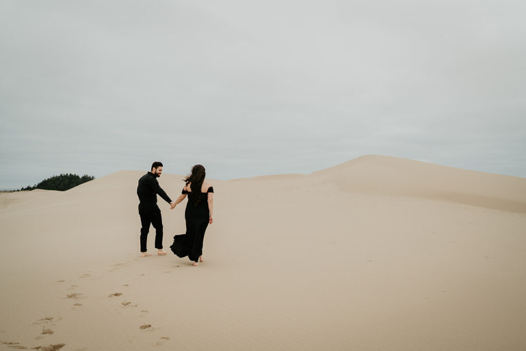 A couple holding hands and wearing black clothes while walking on the Oregon Coast Sand Dunes during their engagement session. 