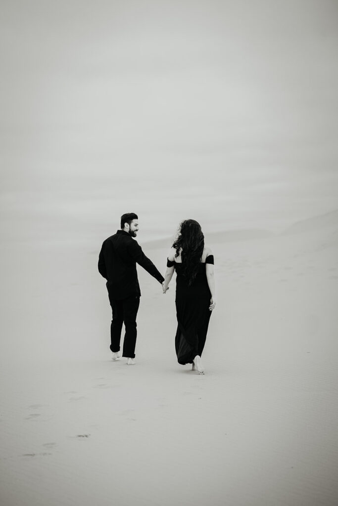 A black and white photo of a couple holding hands and wearing black clothes while walking on the Oregon Coast Sand Dunes during their engagement session. 
