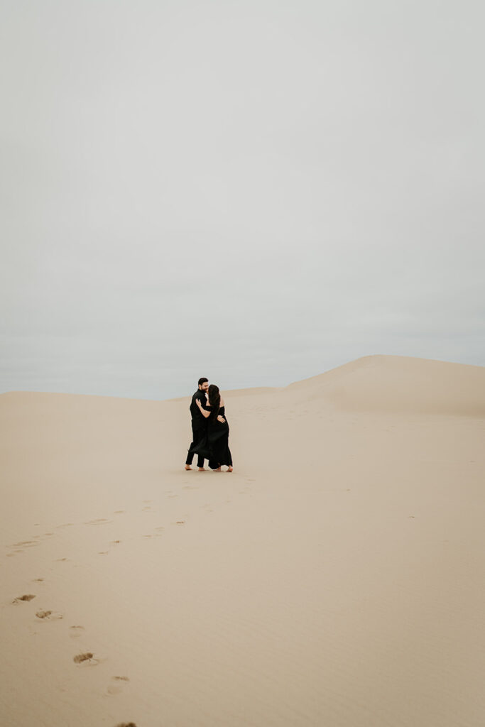 A couple kissing while walking barefoot on the Oregon Coast Sand Dunes and wearing al black. 