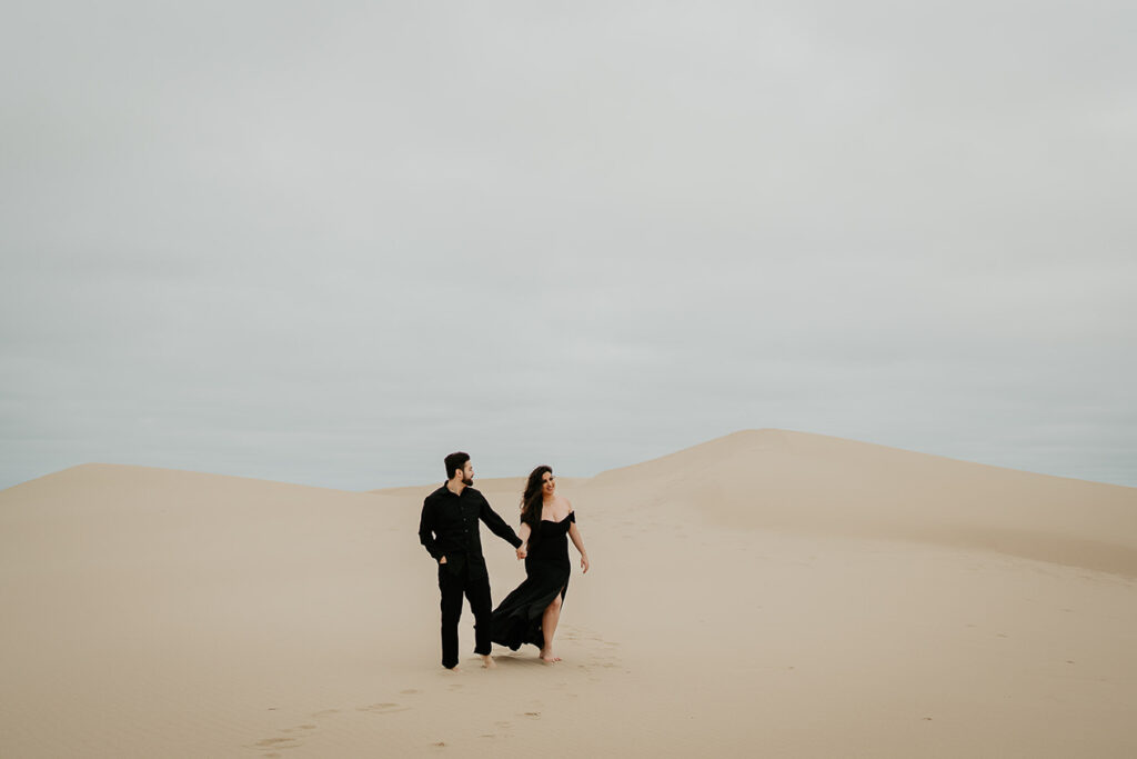 A couple holding hands while walking barefoot on the Oregon Coast Sand Dunes and wearing al black. 