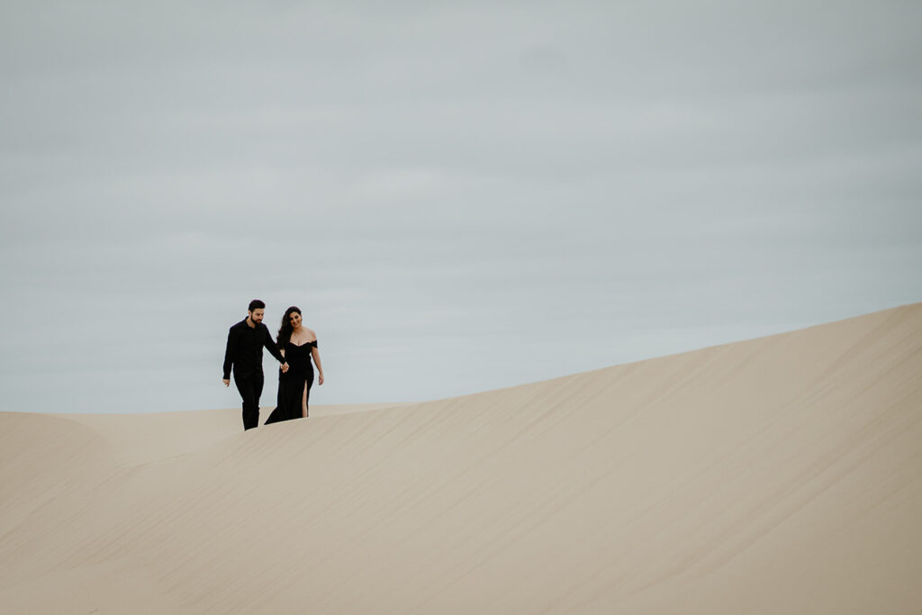 A couple wearing all black and holding hands while walking on a sand dune in Oregon. 