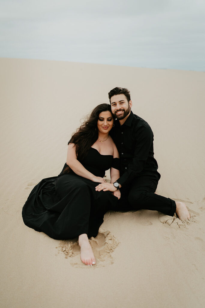 A couple dressing in all black and smiling surrounded by sand dunes. 