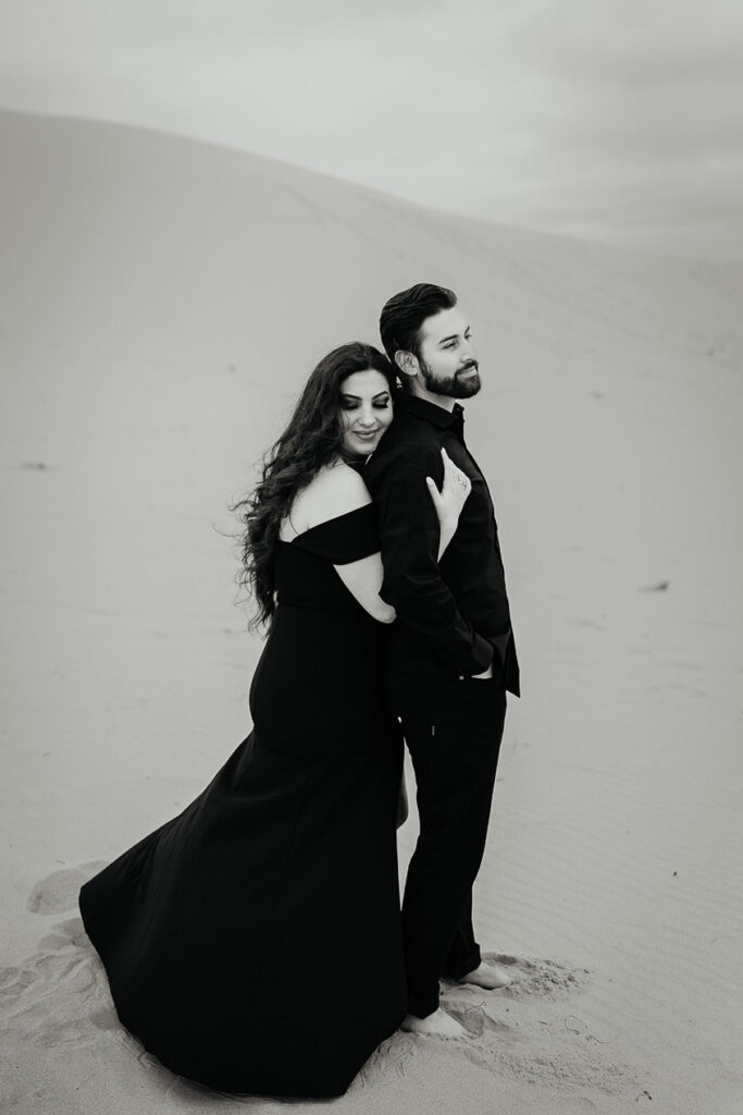 A black and white image of a fiance holding her boyfriend while standing on a sand dune in Oregon during their engagement shoot. 