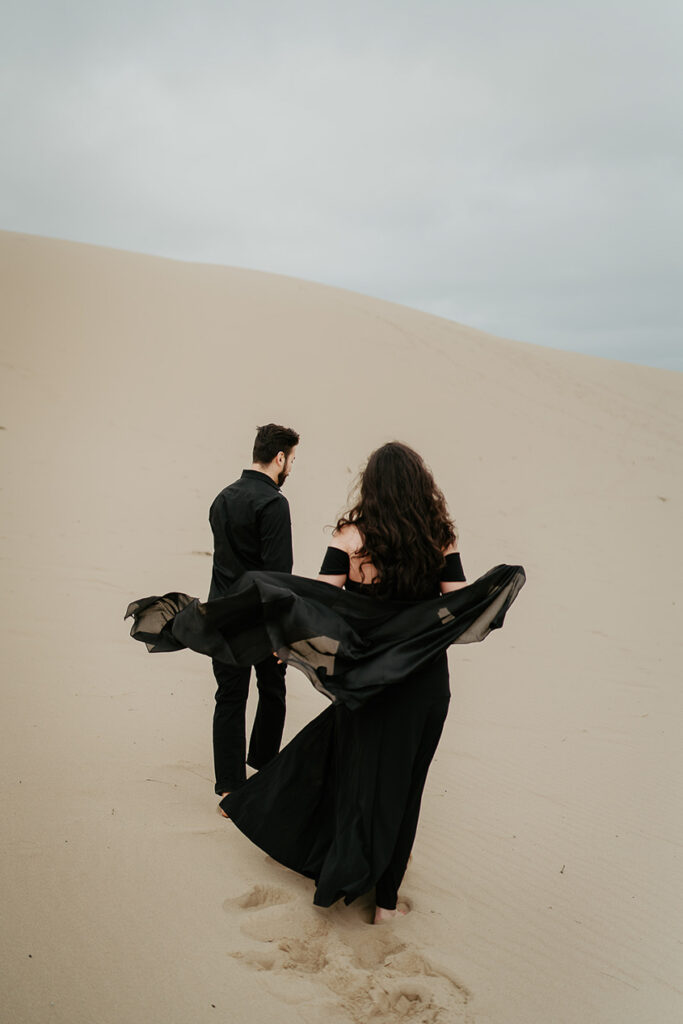 A couple, one with a black dress flowing in the wind and the other in a black button up and pants, walking along the Oregon Coast Sand Dunes during their engagement session. 