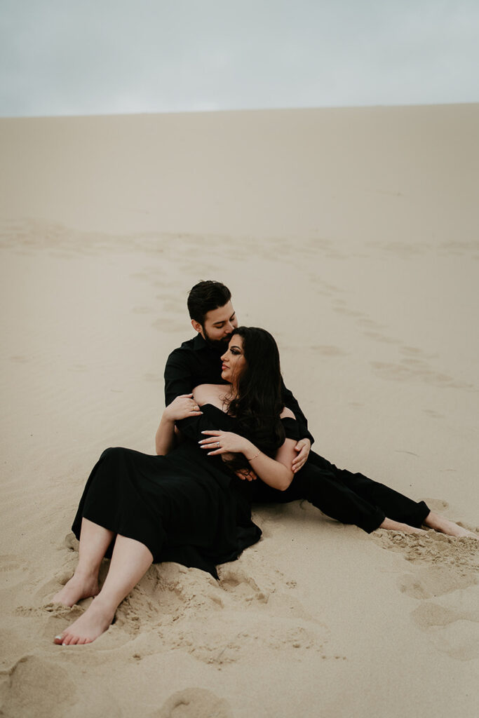 A couple dressed in all black laying in the sand and holding eachother. 