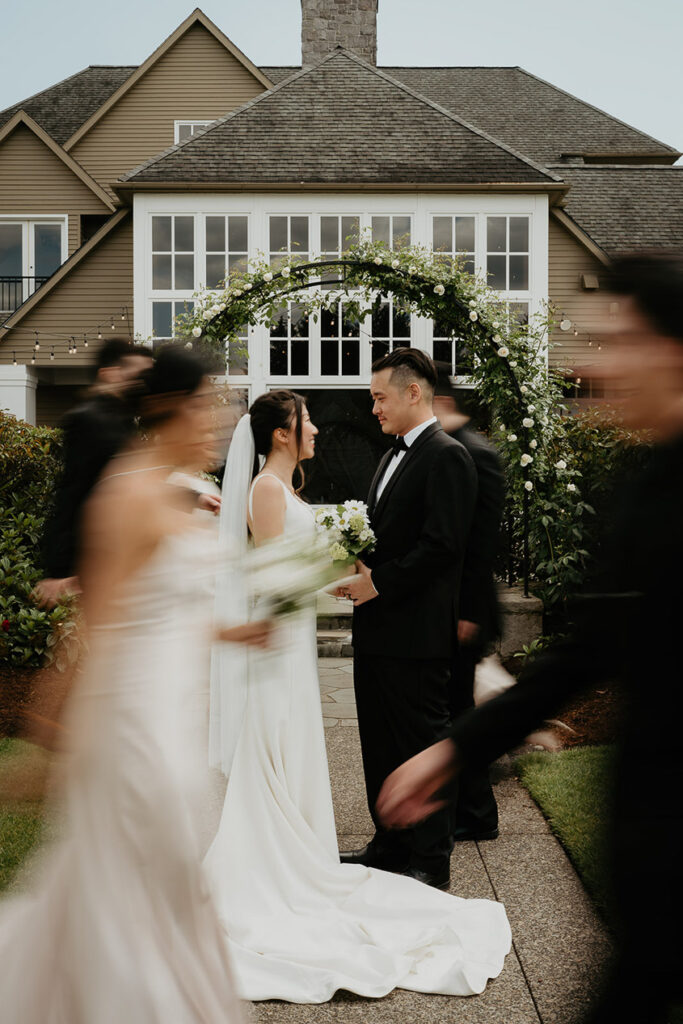 Judith and Joshua holding each other under a flowery, green arch with the blurry wedding party walking past. 