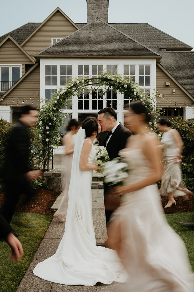 Judith and Joshua kissing under a flowery, green arch with the blurry wedding party walking past. 