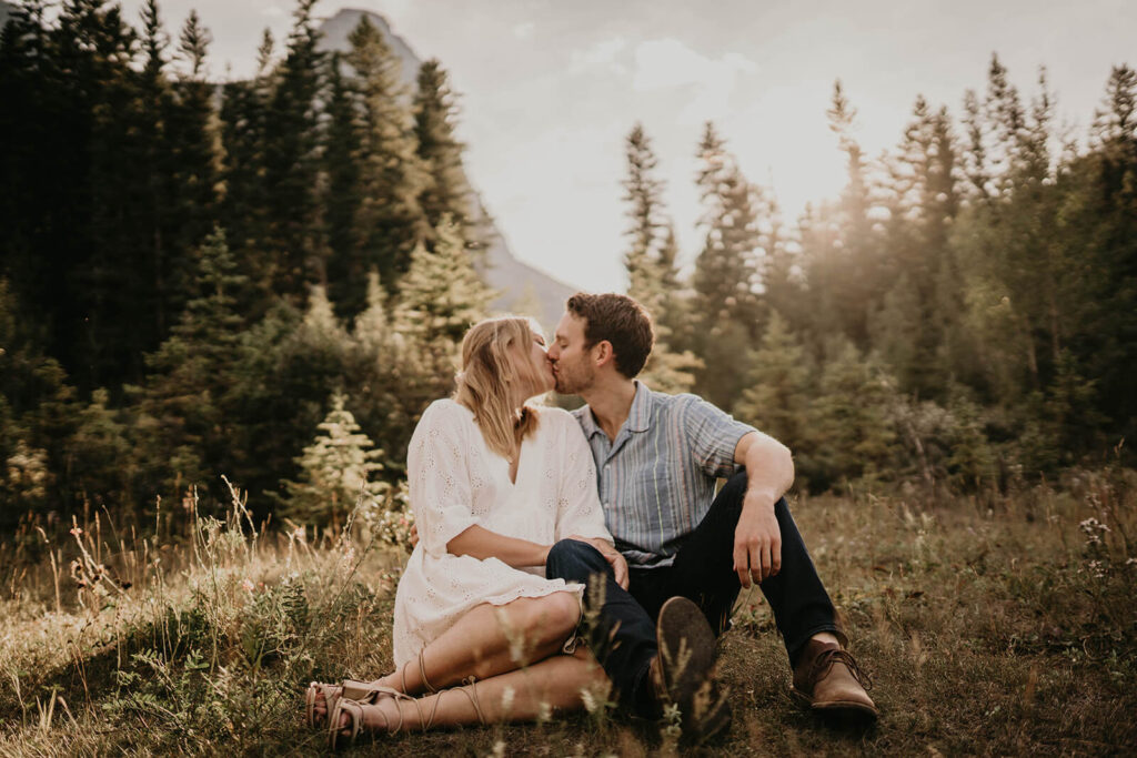 A couple kissing as the sun sets with pine trees and mountains in the background. 