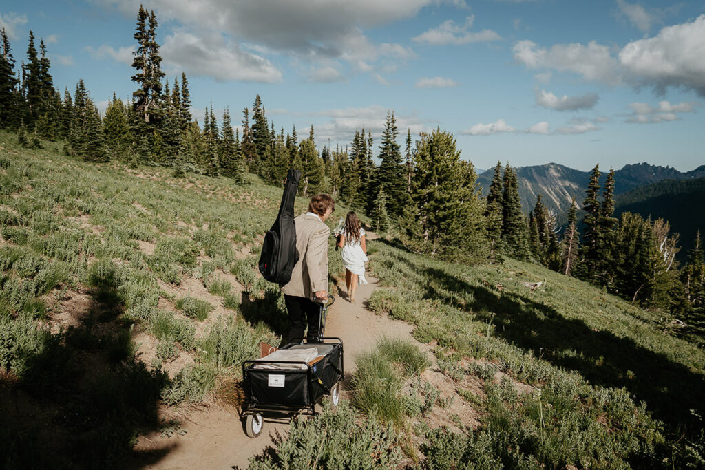 The couple walking along a trail, with the husband holding a guitar and wagon with picnic items. 