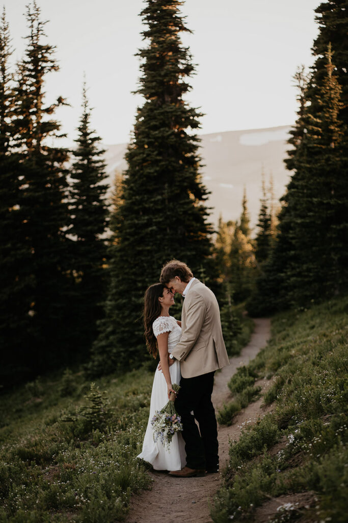 The husband and bride hugging each other on a trail with pine trees in the background at sunset. 
